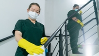 A woman standing in a stairway wiping down the railing with a cloth. She is wearing rubber gloves and a medical mask. 
